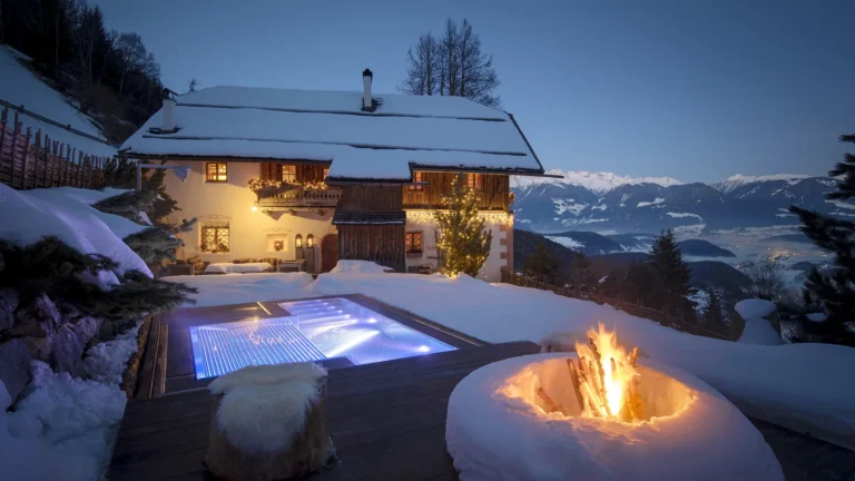 White Deer San Lorenzo Mountain Lodge at dusk, surrounded by snow-covered hills, featuring a glowing fire pit and illuminated outdoor hot tub, offering a serene winter escape with breathtaking views of snow-capped mountains.