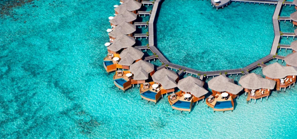 An overwater villa at Baros Maldives sits above crystal-clear turquoise waters, offering a secluded, romantic retreat perfect for Valentine's Day. The serene setting, with a sunset backdrop, creates an idyllic escape for couples.